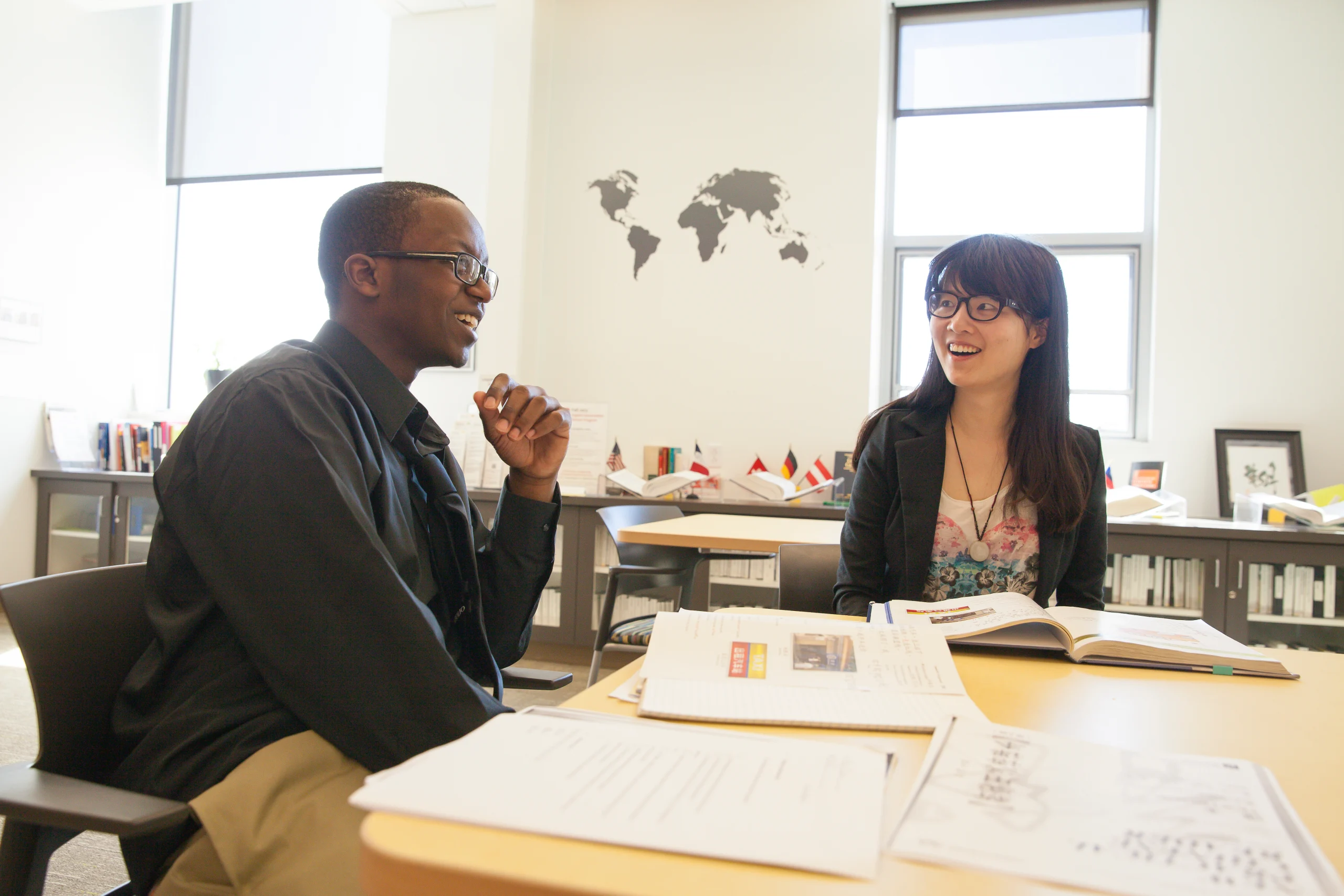Two students sit at a table with books, smiling and talking in a bright classroom with world maps on the wall, discussing English Linguistics and their passion for English teaching.