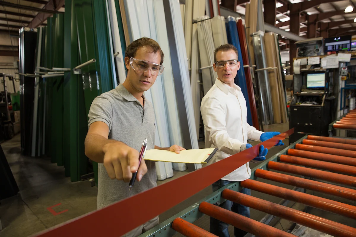 Two men in safety glasses inspect a metal sheet in a factory, one holding a clipboard and pen, demonstrating an Integrated Business approach to engineering processes.