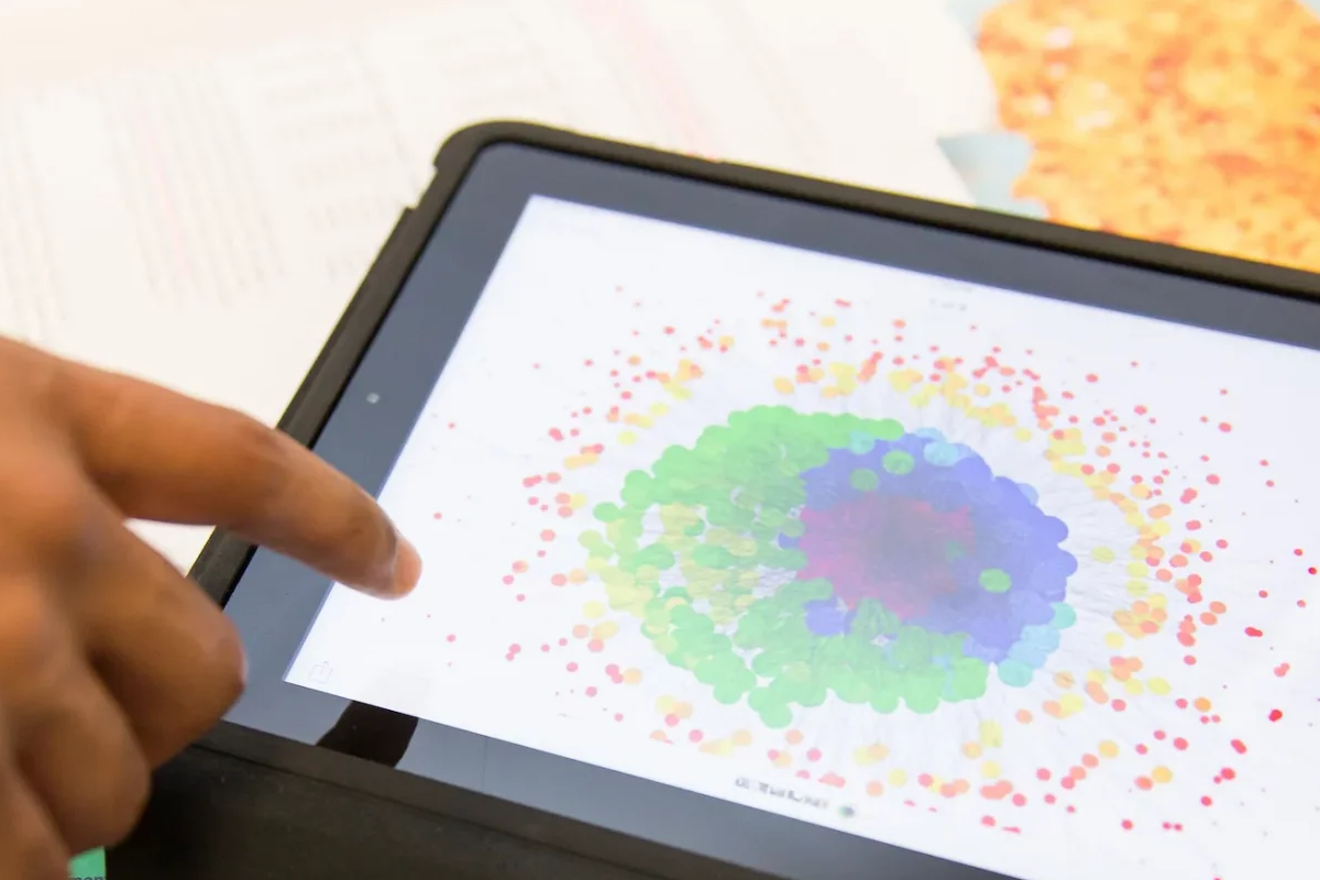 A hand points at a tablet displaying a colorful Business Analytics data visualization with clustered dots in green, purple, yellow, and red.
