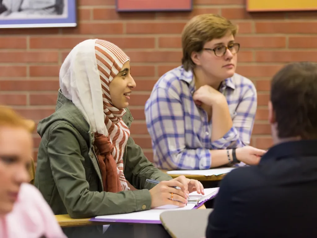 Two students sit at desks in a classroom, attentively listening and taking notes.