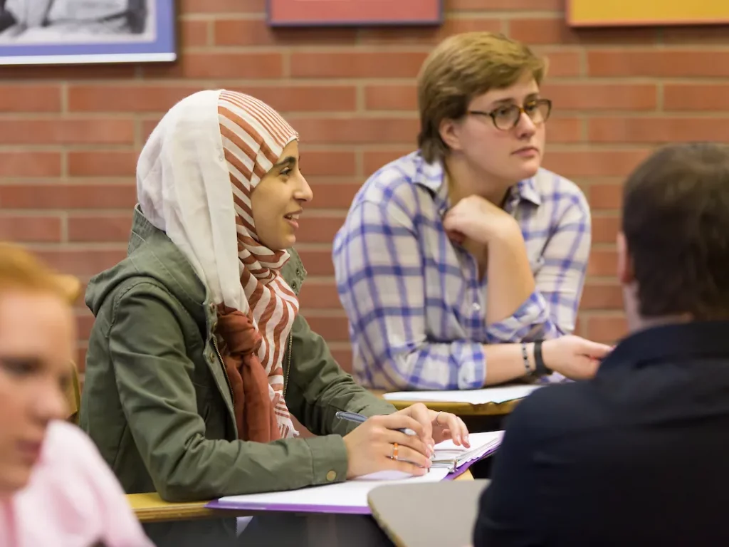 Two students sit at desks in a classroom, one wearing a hijab, both listening and taking notes during their Social Work Masters program.