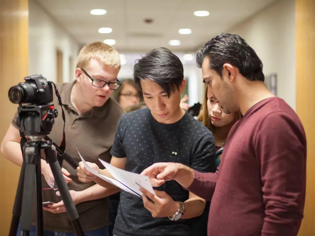 Three people stand in a hallway, reviewing papers together beside a camera on a tripod, suggesting a collaborative Cinema or Media Studies project.