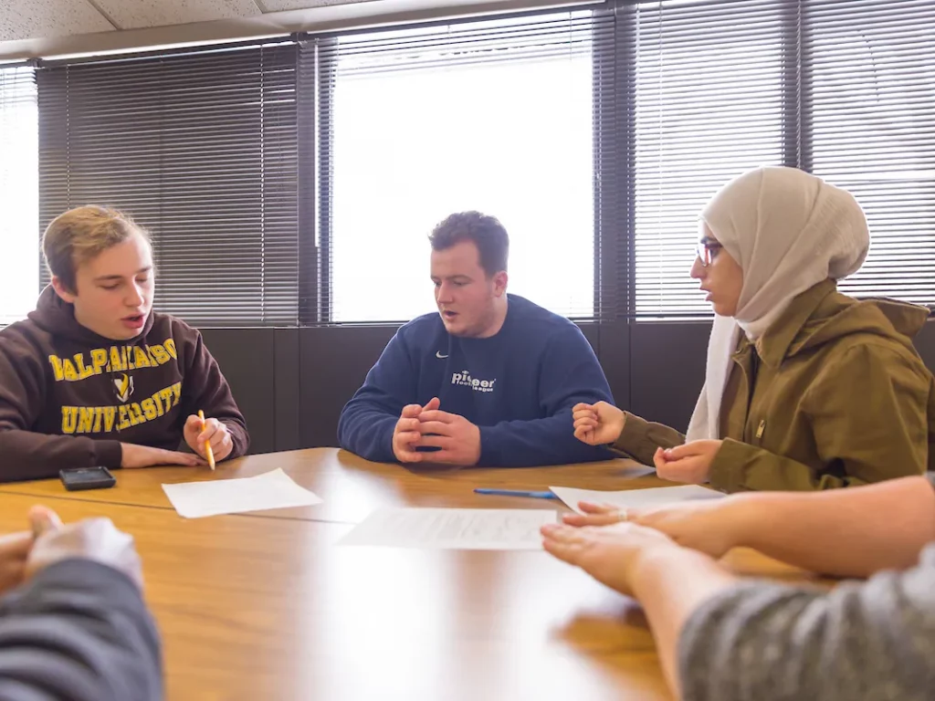 Three students sitting at a table discussing papers, one wearing a hijab, in a bright room.