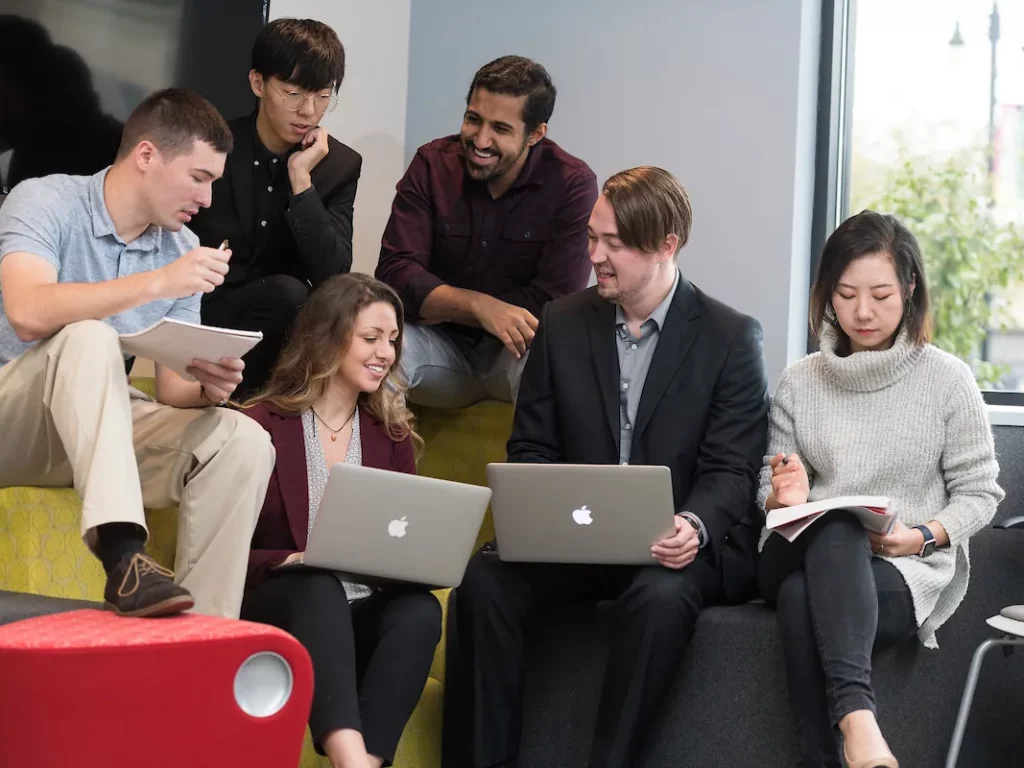 Six people sit together in a modern office space, some using laptops and notepads, engaged in discussion and collaboration for a Business Administration project.