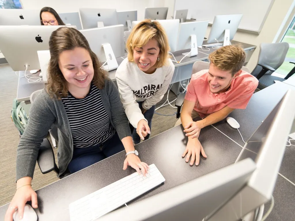 Three students smiling and working together at desktop computers in a modern classroom while collaborating on a public health degree project.