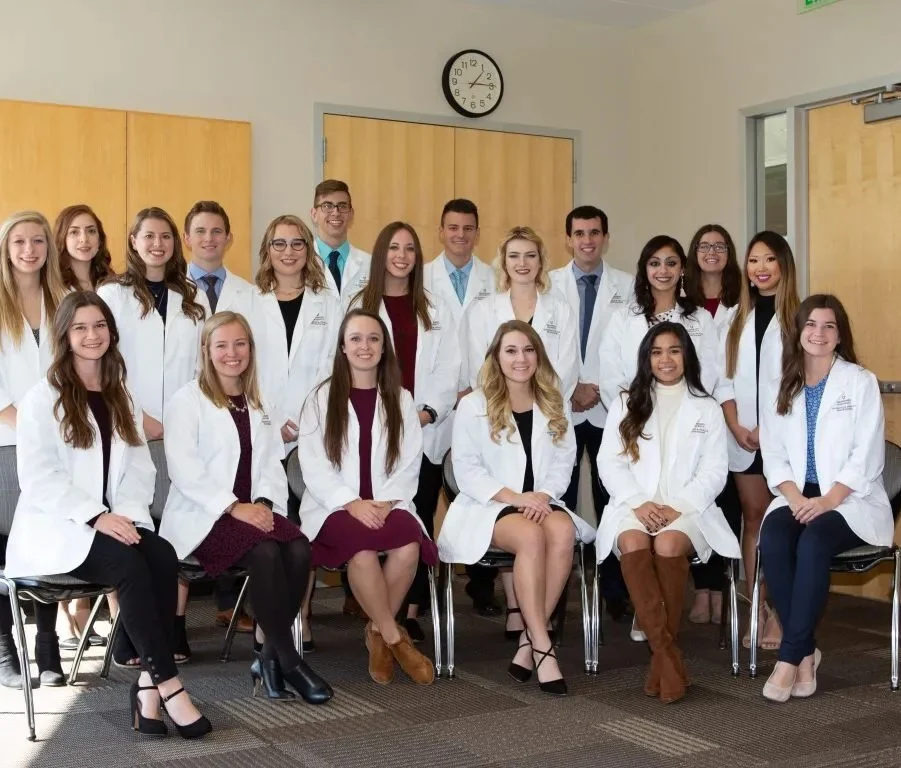 A group of Health Science students in white coats pose for a photo in a classroom, smiling at the camera.