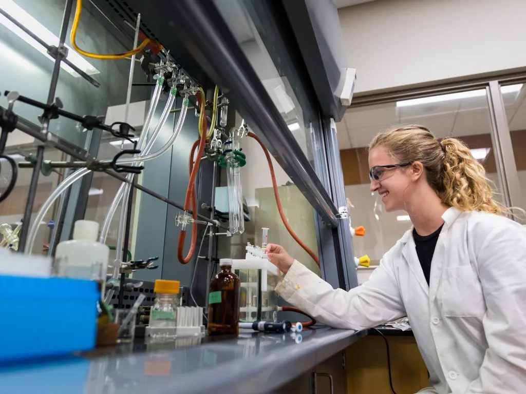 A scientist in a lab coat works at a laboratory bench with biochemistry equipment and bottles.