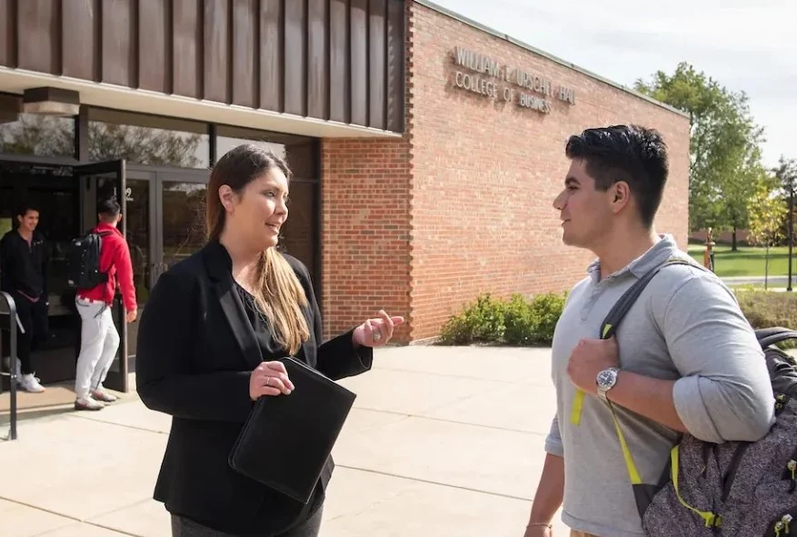 Two students talk outside a college building on a sunny day, one holding a folder, the other a backpack.