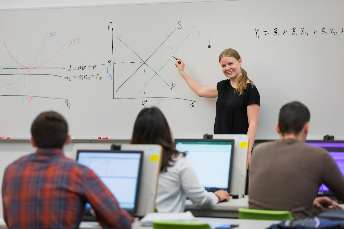 A woman explains supply and demand graphs on a whiteboard to students using computers in a Business Economics Certificate classroom.