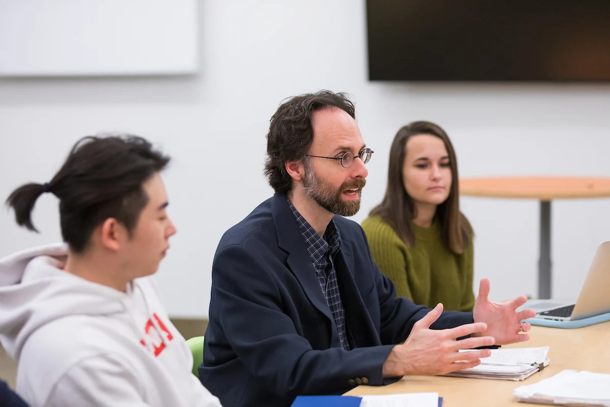 A man speaks at a table with two students, discussing papers and a laptop in a classroom setting, as they delve into topics from the Masters in International Economics and Finance program.
