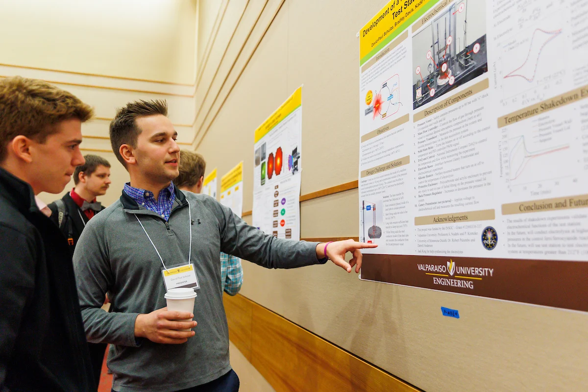 Two men discuss an engineering research poster at a university event, one pointing at the display and actively sharing your research findings.