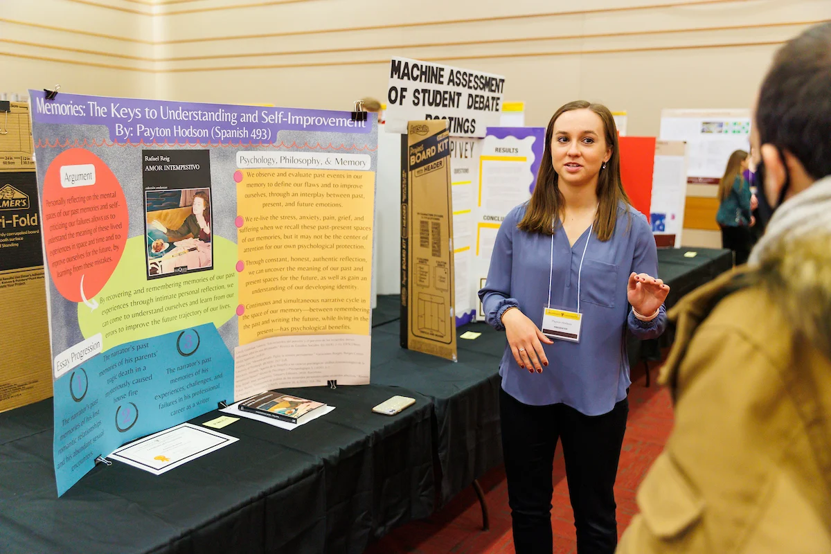 A student presents a colorful research poster at the Symposium on Undergraduate Research and Creative Expression, engaging an attendee in discussion.