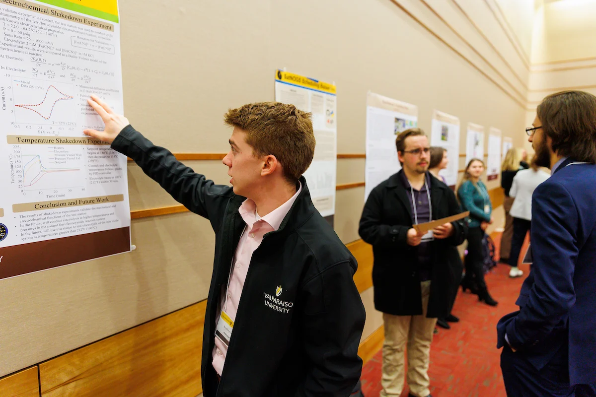 A young man is sharing his research as he presents a scientific poster to attendees at a conference in a hallway.