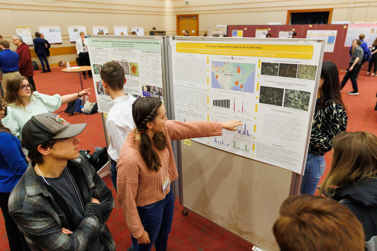 Students share their research and present posters at a science fair in a large indoor room.