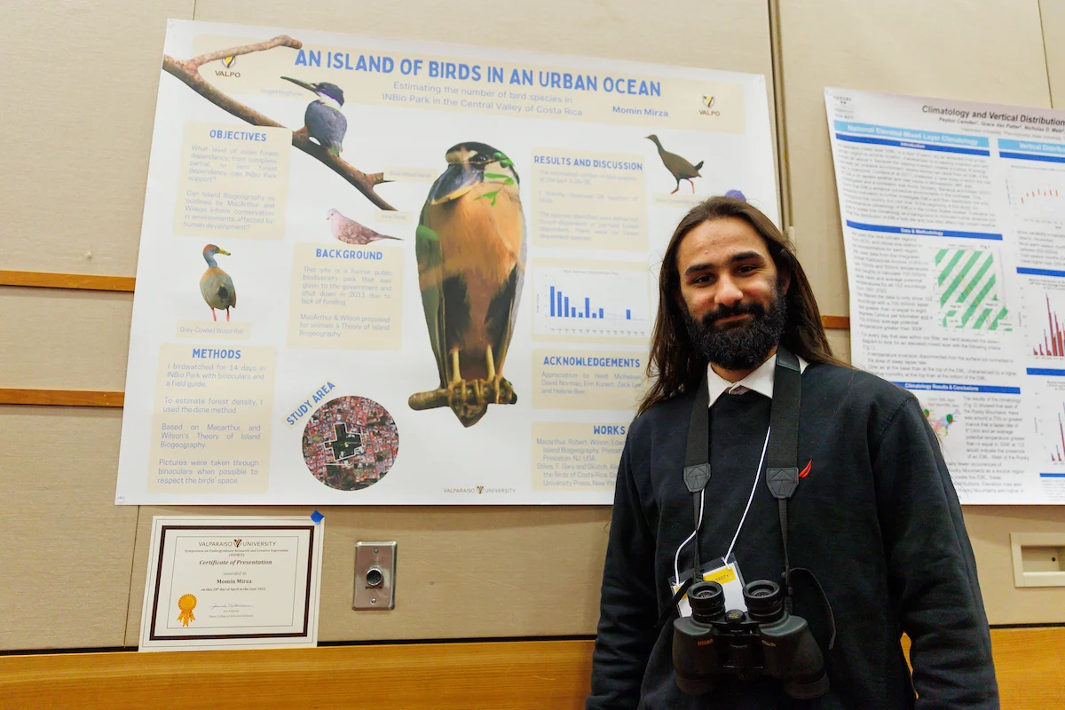 Man with binoculars stands in front of a scientific poster about urban birds at the Symposium on Undergraduate Research and Creative Expression.