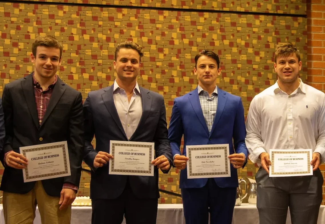 Four young men in formal attire stand indoors, each proudly holding a "College of Business" certificate, having completed the Entrepreneurship Minor program, against a patterned wall.