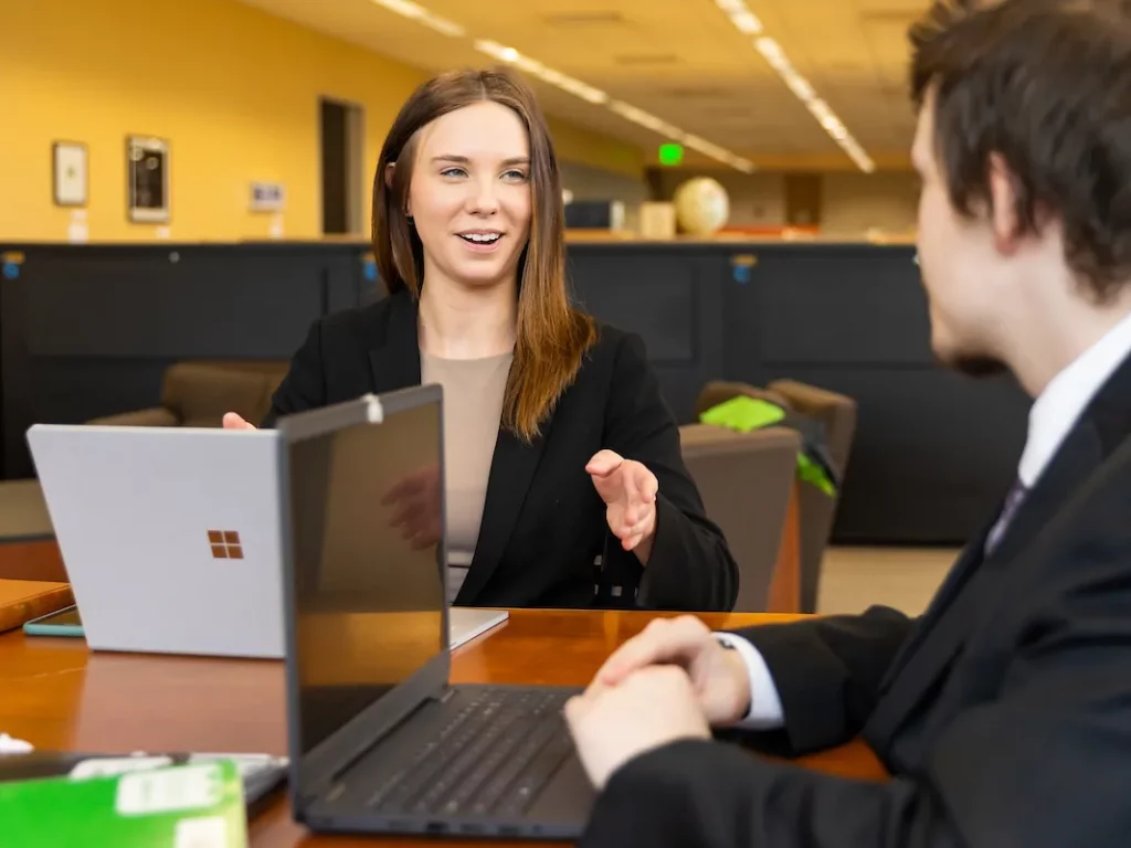 Two people in business attire sit at a table with laptops, engaged in conversation in an office setting.