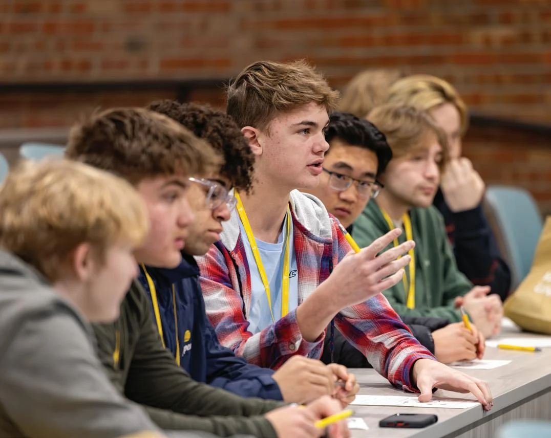 Several students sit in a classroom, taking notes and listening attentively to a lesson on business fundamentals; one student in a plaid shirt speaks or asks a question, holding a pencil.