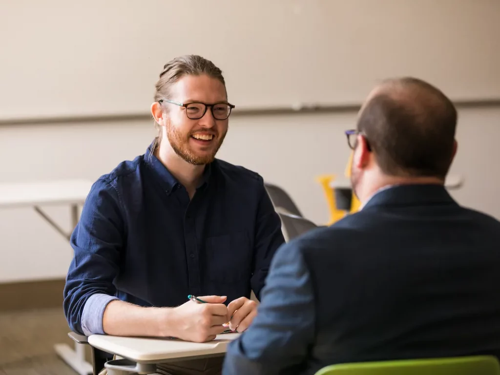 Two men sit across from each other at a desk, having a friendly conversation in a classroom setting, discussing topics related to Masters in Clinical Mental Health Counseling.