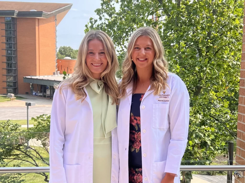 Two women in white lab coats, pursuing Health Science, smile on a balcony with trees and a brick building in the background.