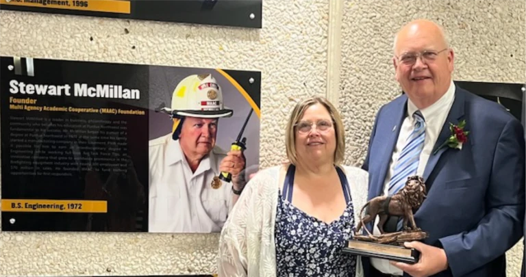 Two people stand smiling; the man holds a lion statue trophy by a plaque featuring Stewart McMillan, celebrating their achievement at the Innovation Hub.