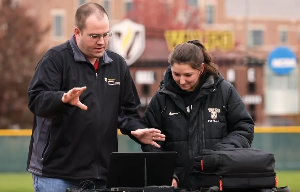 Two adults stand outdoors at a table with a laptop, discussing bioengineering. One gestures while the other looks at the screen. Both wear jackets, with a sports field and buildings in the background.