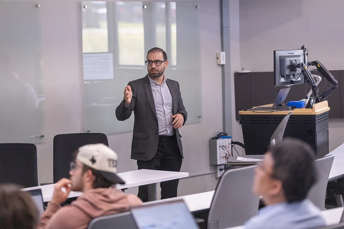 A man in a suit gestures while speaking to MBA students in a modern classroom with computers, discussing Business Administration concepts.