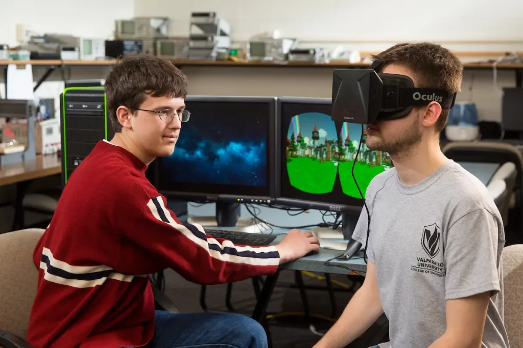 Two young men explore a virtual reality headset and computer in a technology lab, highlighting the innovative world of computer engineering.