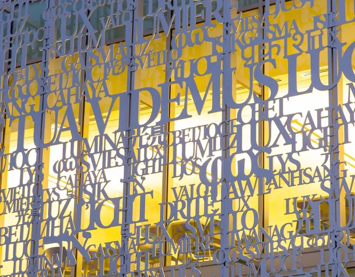 White cut-out letters in various languages cover a glass wall, with bright yellow light shining through—evoking the interconnected pathways of neuroscience and communication.