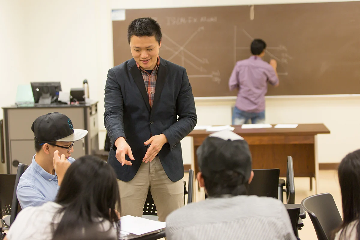 A teacher discusses concepts of Applied Econometrics with students while another person writes on a chalkboard in a classroom.