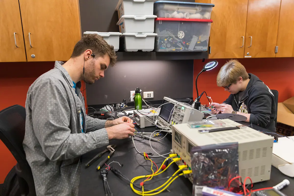 Two people work on electronics projects at a cluttered desk with tools, wires, and equipment, showcasing hands-on learning in Computer Engineering and Electrical Engineering.