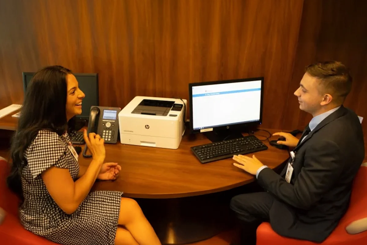 Two professionals in business attire smile and converse at a desk with a computer, printer, and phone, reflecting the collaborative spirit found in Master of Healthcare Administration (MHCA) programs.