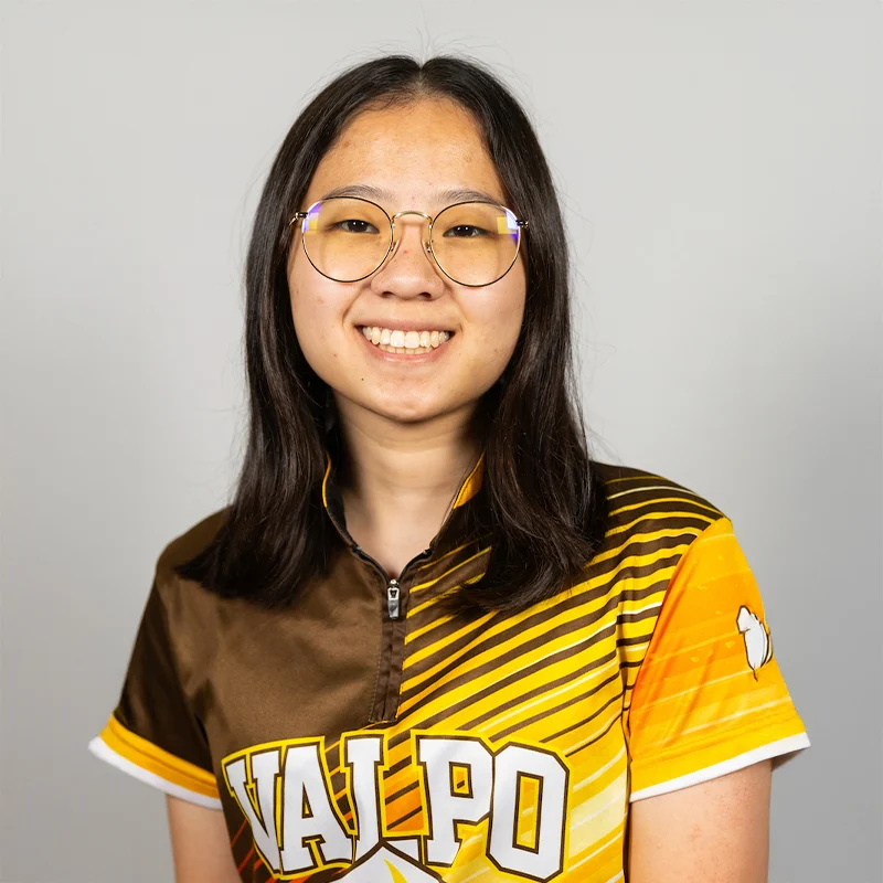 Smiling person with glasses, Lilu Smith, wearing a yellow and brown "VALPO" sports shirt, poses confidently against a plain background, reflecting a passion for Computer Science.