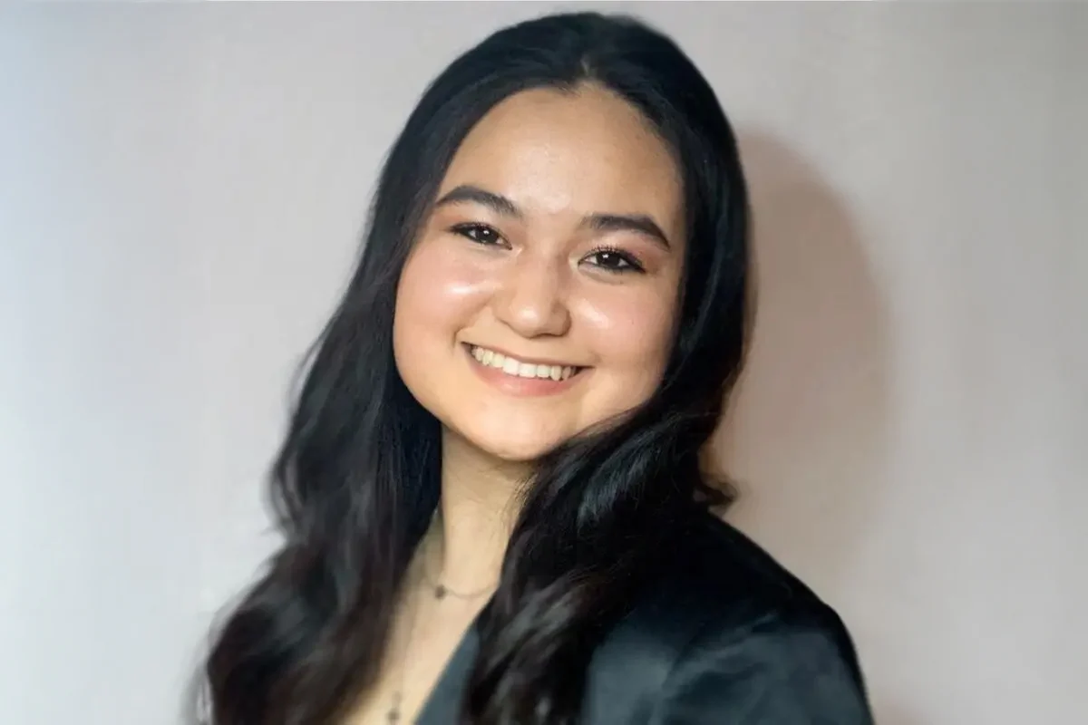 A young woman with long, dark hair smiles at the camera against a plain light background, capturing a moment of simple, natural art.