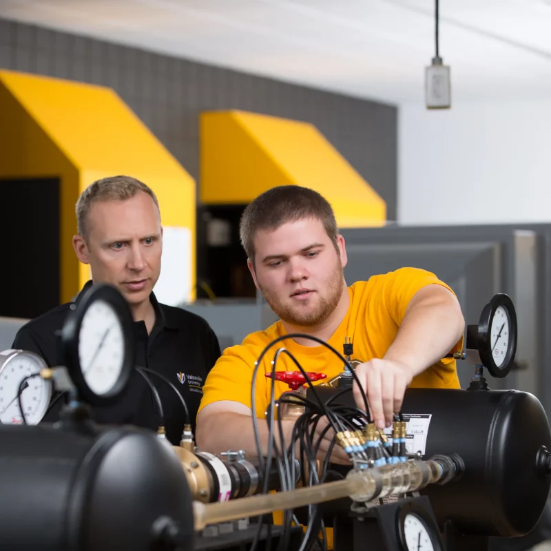 Two men work with pressure gauges and machinery in a Mechanical Engineering lab, one explaining concepts as the other adjusts controls.