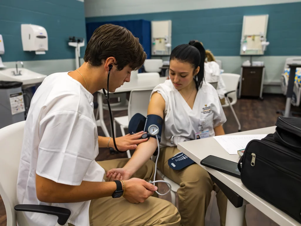 Two nursing students practice taking blood pressure in a classroom with medical equipment in the background.