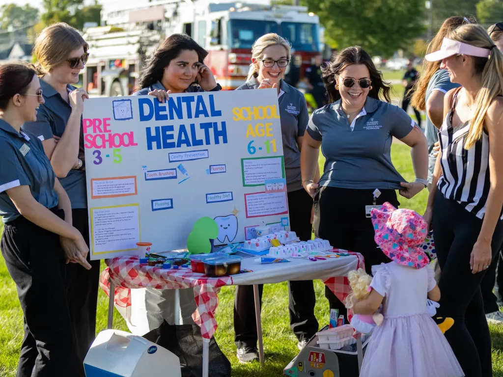 A group of women promotes public health by showing a dental health board to a young child at an outdoor event on a sunny day.