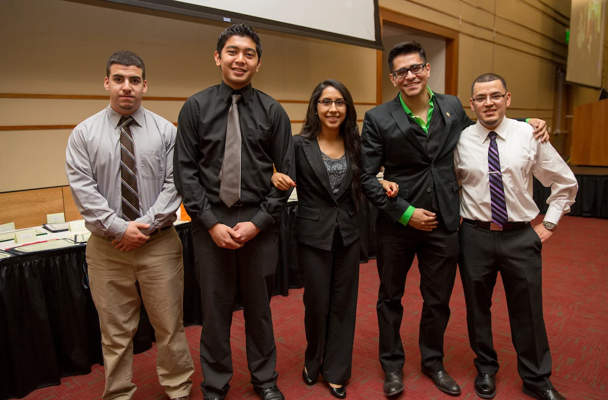 Five people in business attire stand together, smiling, in a conference or banquet room with red carpet—reflecting the inclusive spirit of multicultural organizations.