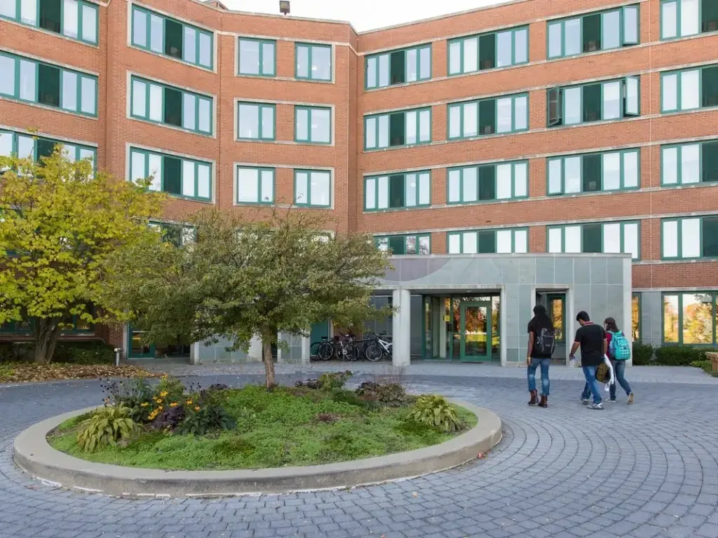 Four-story brick dorm building, Wehrenberg Hall, with green windows; three people walk toward the entrance, trees and bikes nearby.