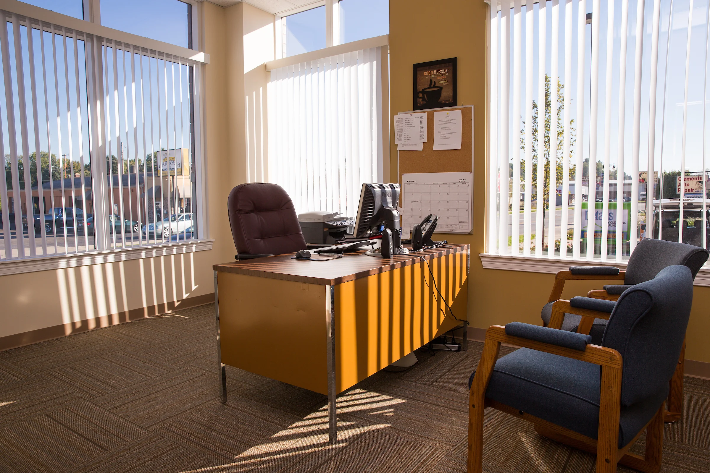 Sunlit office with a desk, chair, computer, and two guest chairs by large windows with vertical blinds, creating a bright atmosphere that promotes wellness and health.