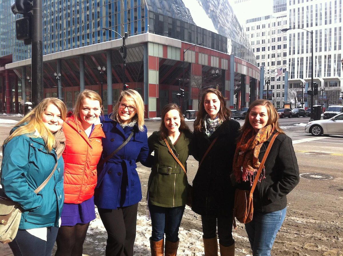 Six women in winter coats smile on a sunny city street with modern buildings in the background, embodying the spirit of Uplift Valpo.