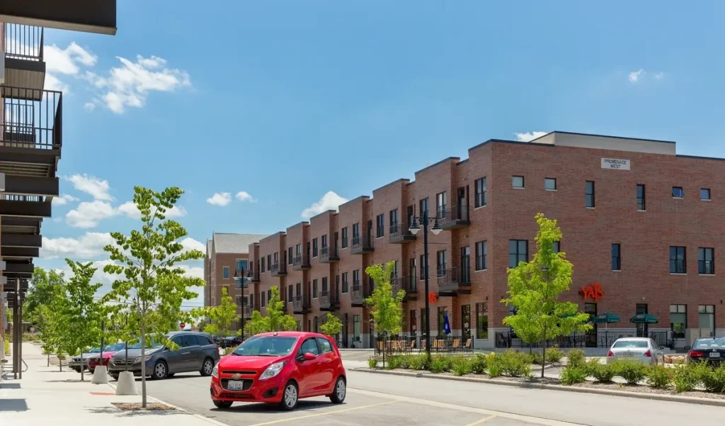A red car parked on a street lined with modern brick Promenade Apartments and young trees under a blue sky.