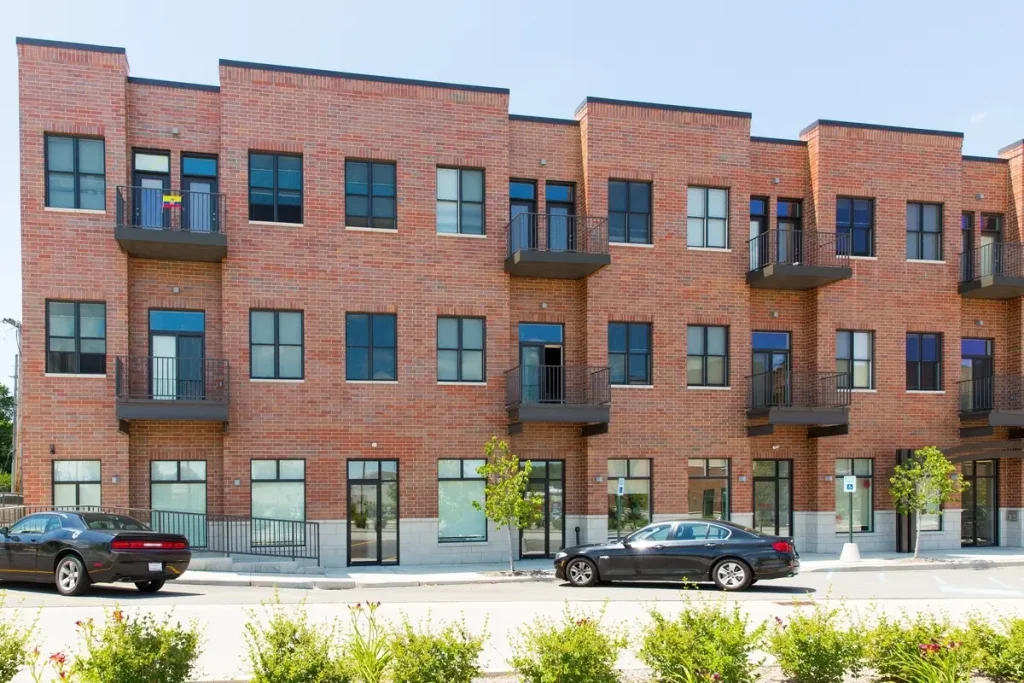 Promenade Apartments is a three-story modern brick building featuring balconies, large windows, and parked cars in front.