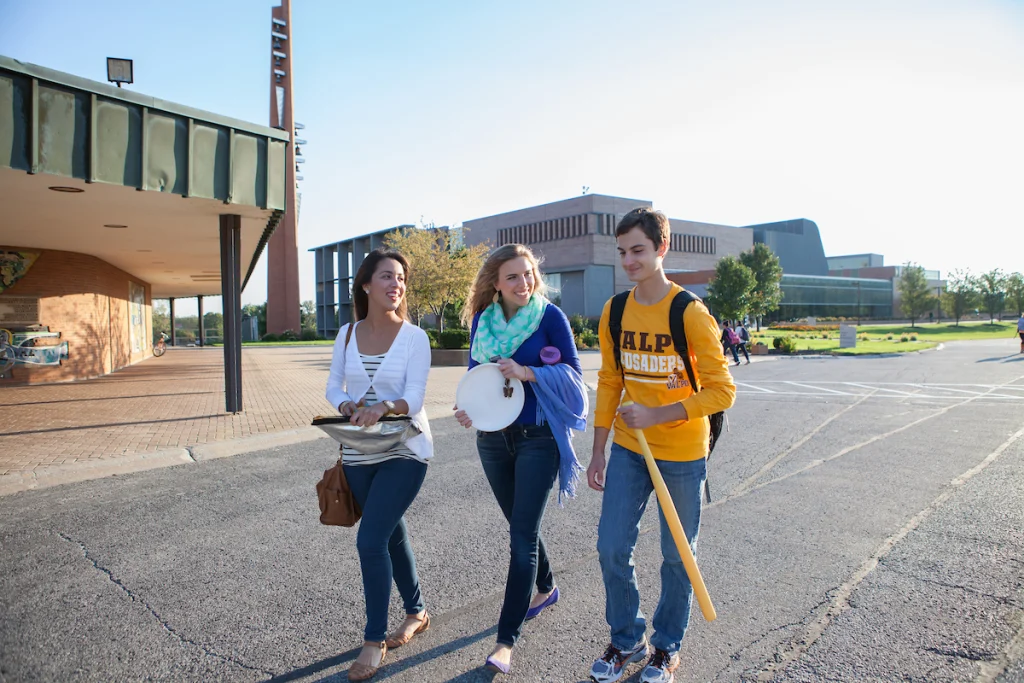 Three smiling students walk outside the Hesse Learning Resource Center, carrying musical instruments and talking together on a sunny day.