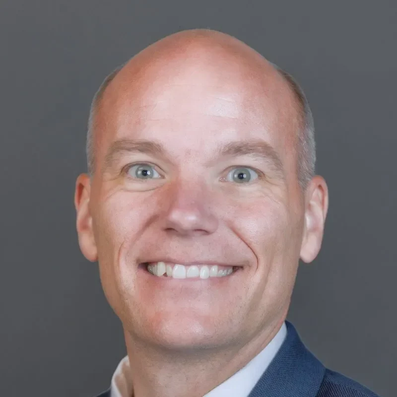Smiling bald man in a suit jacket, exuding confidence and professionalism, ideal for communications roles, against a plain gray background.