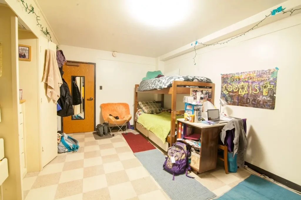 A cozy college dorm room in Guild-Memorial Hall with bunk beds, a desk, backpacks, and string lights brightening the wall.
