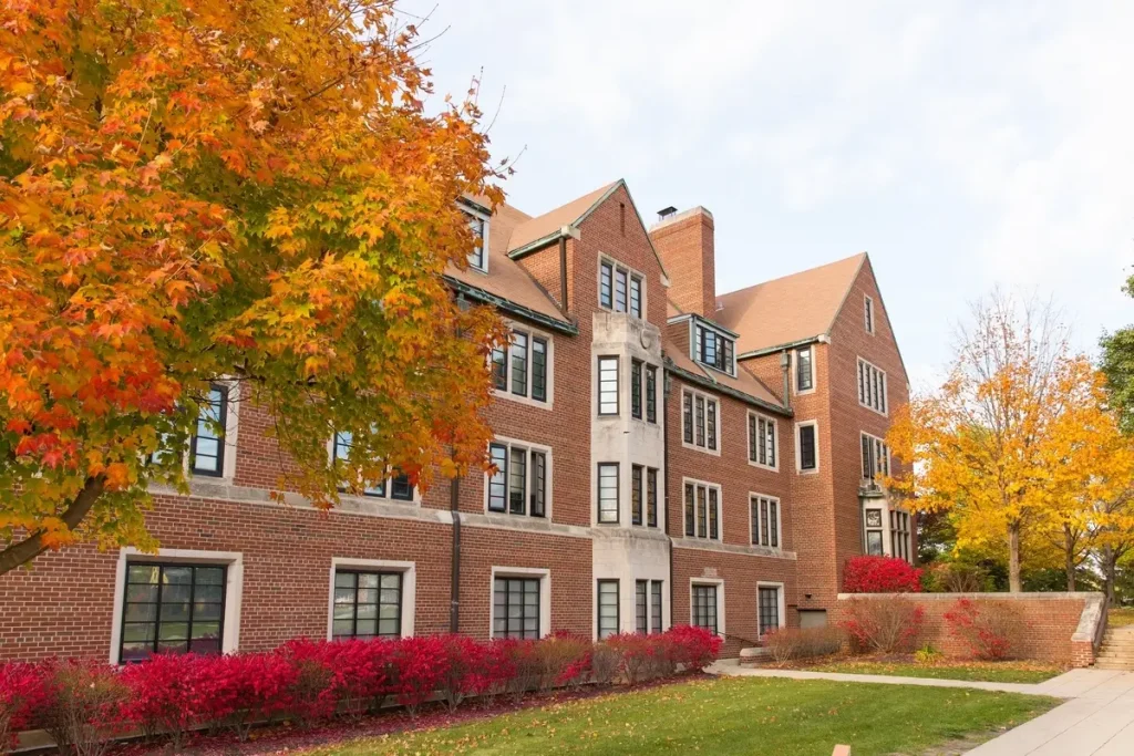 Guild-Memorial Hall stands as a red-brick campus building framed by vibrant fall foliage, red bushes, and a partly cloudy sky.