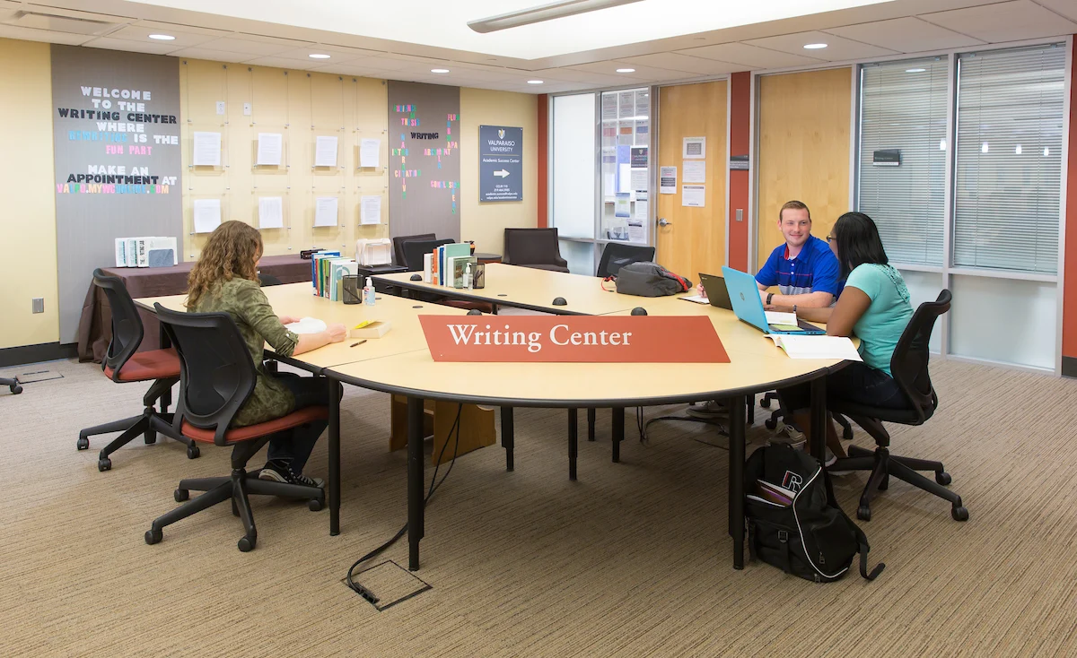 Three students sit at a round table in a Writing Center, working on laptops and papers with the guidance of Writing Consultants.