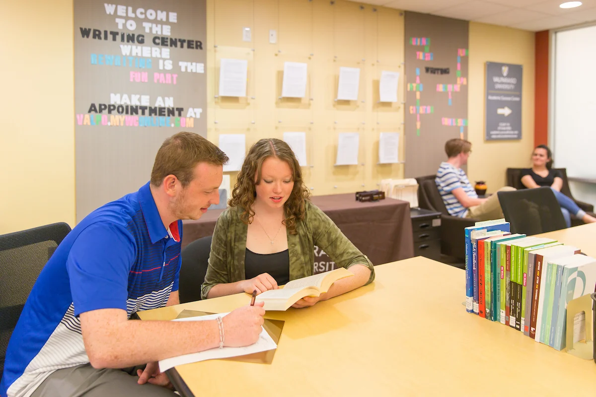Two people sit at a table in a writing center, reading and discussing a book with guidance from Writing Consultants.
