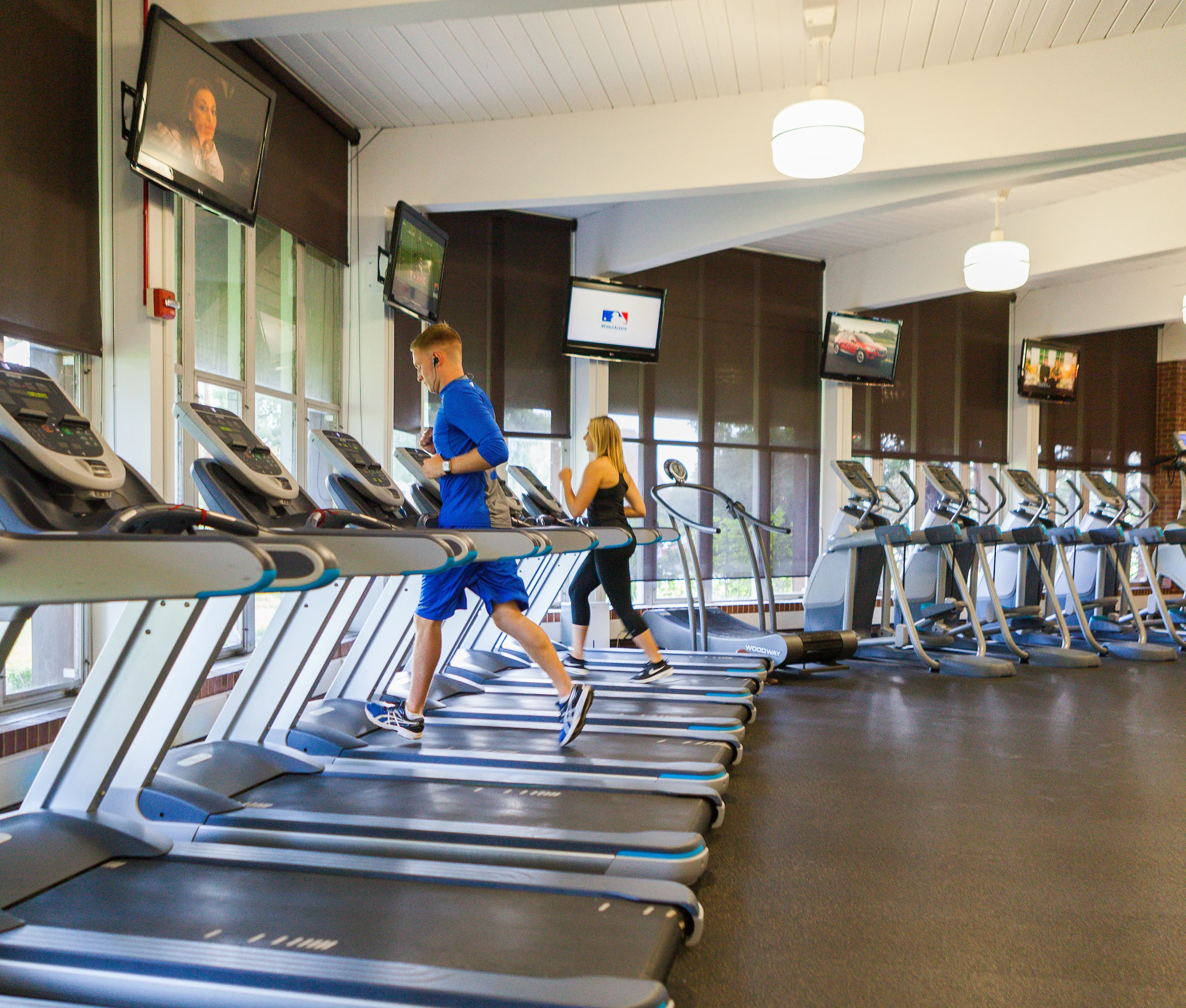 Two people running on treadmills in a gym with several TVs mounted on the walls, embracing fitness for overall health and wellness.